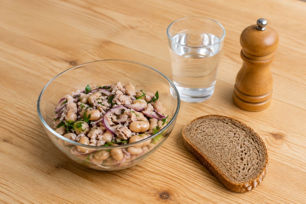 Tuna and white bean salad in a glass bowl with rye bread, a glass of water and a pepper mill on a pine table