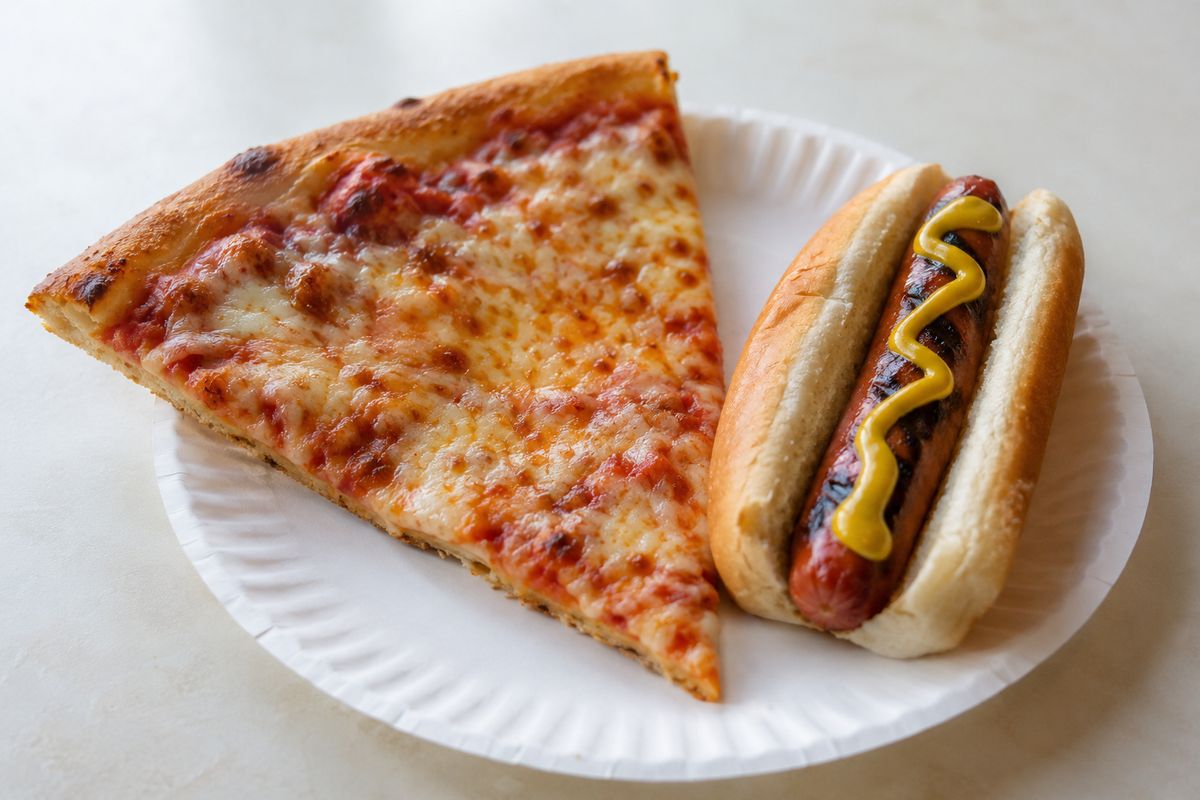 Una porción de pizza de queso en un plato de papel junto a un hot dog en su pan con una línea de mostaza