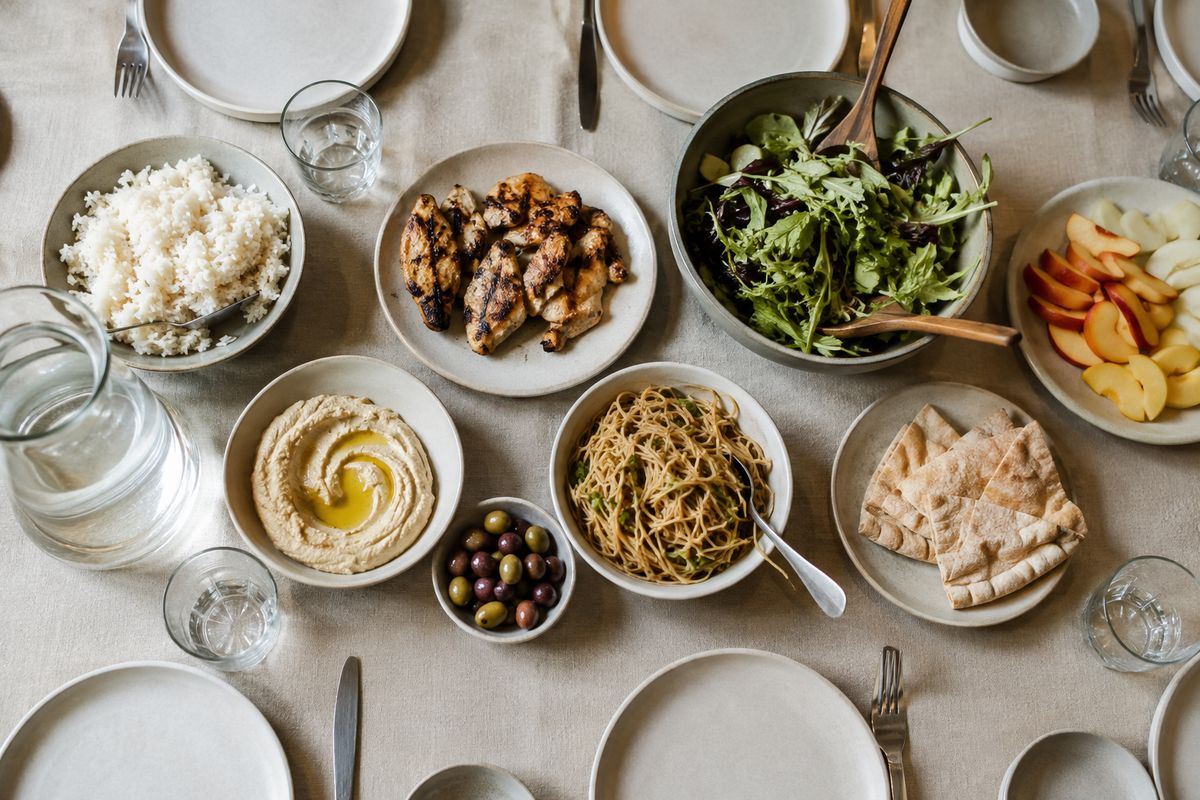Overhead view of a long table with many small dishes: rice, grilled chicken, salad, fruit, noodles, hummus, bread and olives
