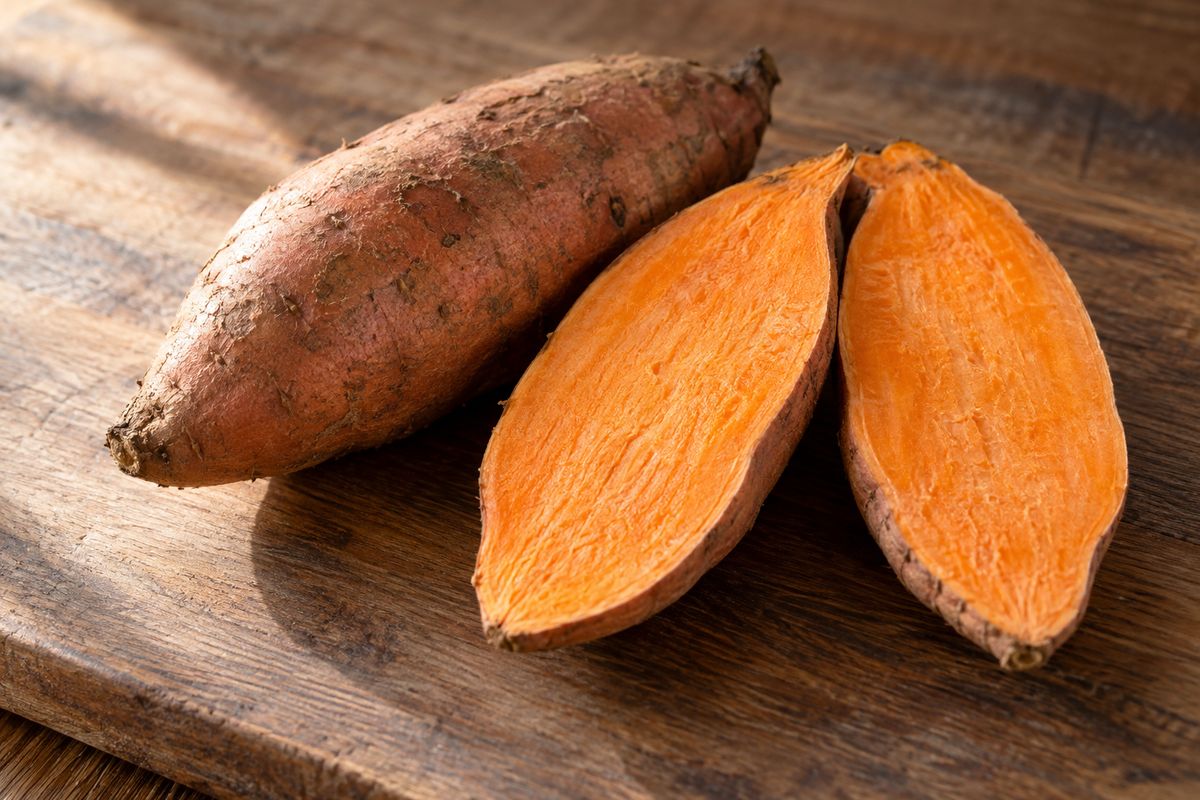 Two whole orange-fleshed sweet potatoes on a wooden board, one cut lengthwise to show the flesh.